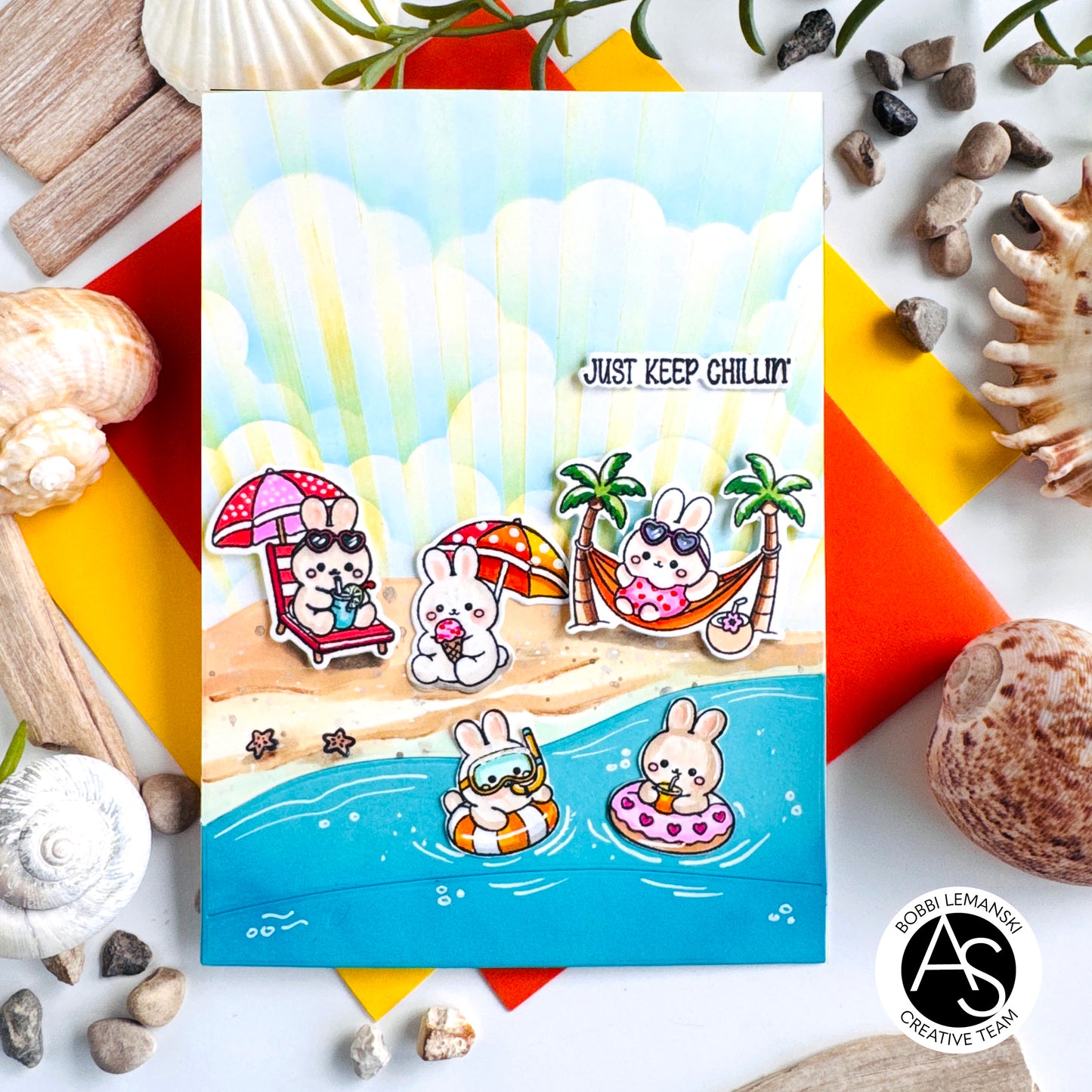 A square handmade greeting card featuring a beach scene with cartoon rabbits relaxing by the shore, surrounded by seashells and colorful paper accents. The card has the text 'JUST KEEP CHILLIN' and a circular 'AS Creative Team' logo in the bottom right corner. Alex Syberia Designs
